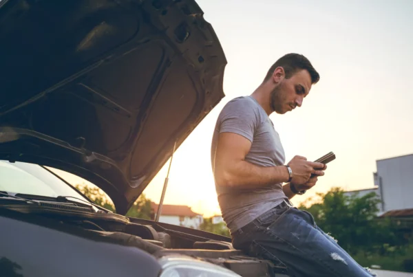 A man leans on the front of a car with the hood open, checking his phone at sunset beside the road.
