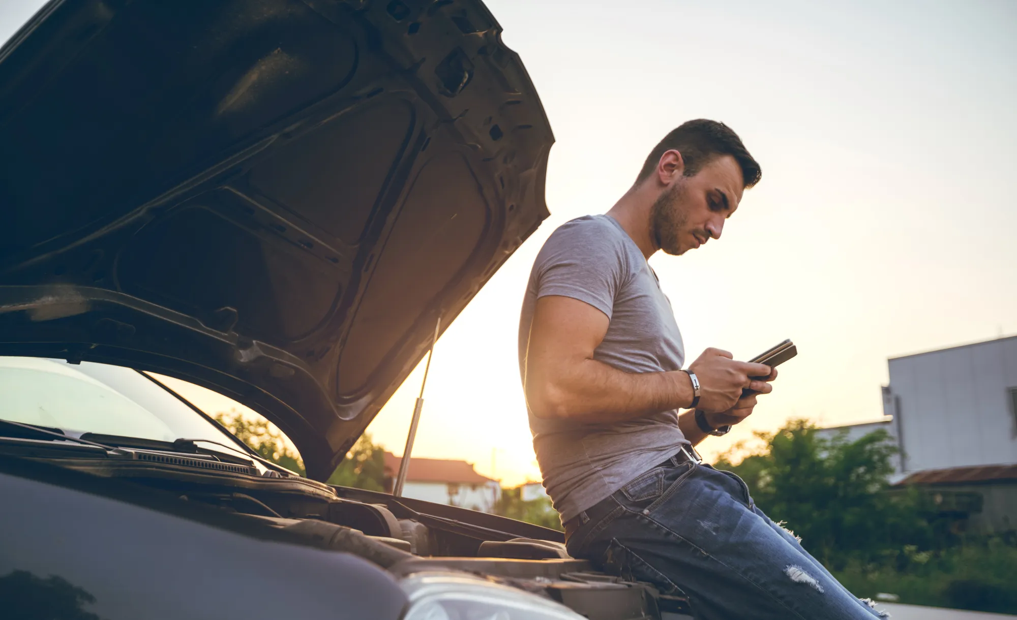A man leans on the front of a car with the hood open, checking his phone at sunset beside the road.