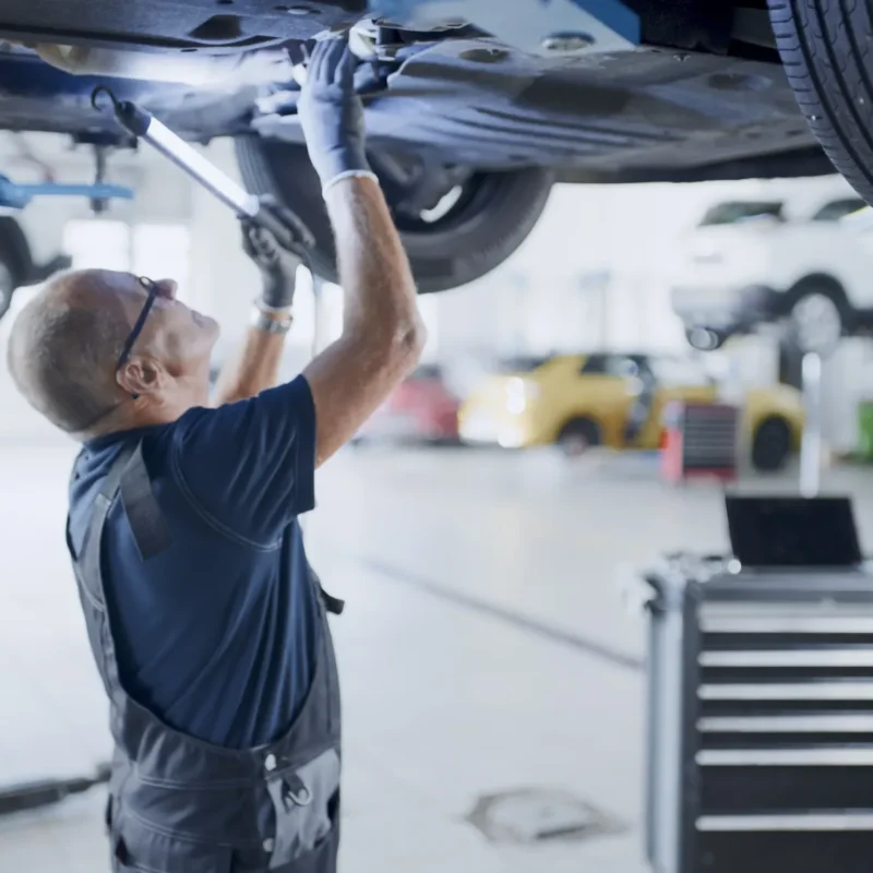 A mechanic in overalls and safety glasses works underneath a car raised on a lift in a busy auto repair shop.