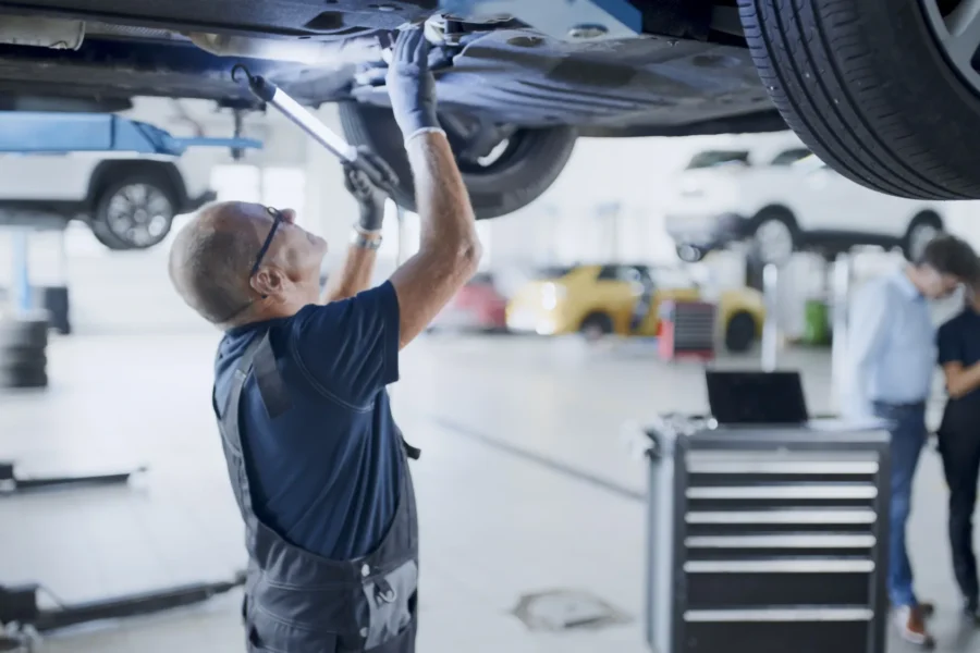 A mechanic in overalls and safety glasses works underneath a car raised on a lift in a busy auto repair shop.