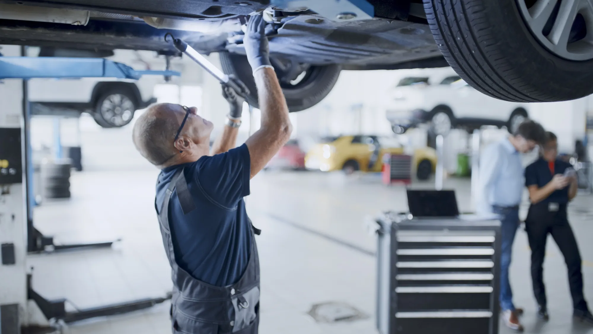 A mechanic in overalls and safety glasses works underneath a car raised on a lift in a busy auto repair shop.