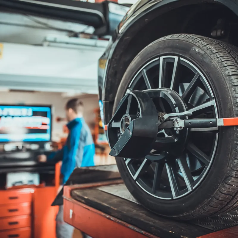 A wheel alignment sensor clamped to the alloy wheel of a car raised on a shop lift, with a technician at the alignment machine in the background.