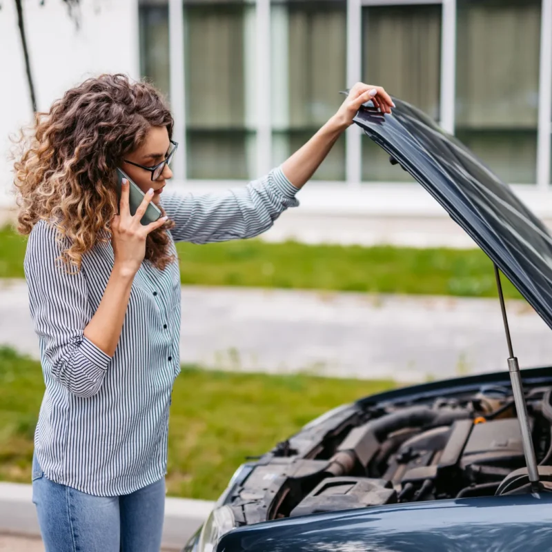 A woman holds open the hood of her car at the roadside while talking on her cell phone.