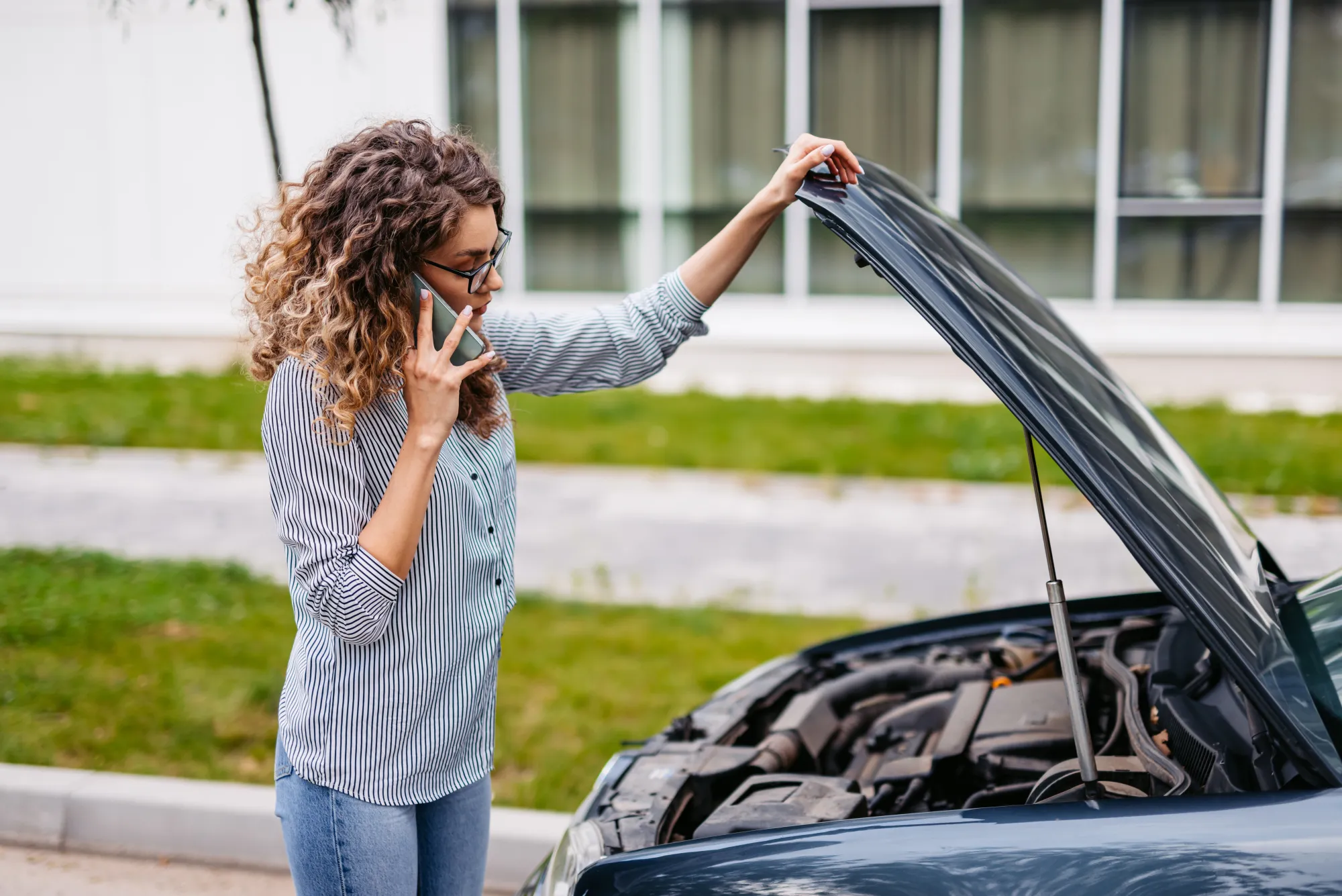 A woman holds open the hood of her car at the roadside while talking on her cell phone.