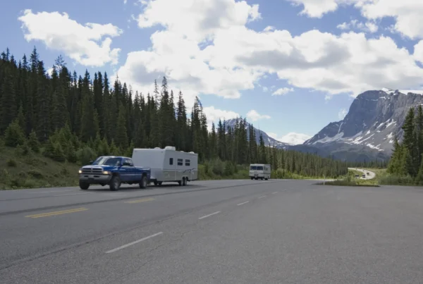 A blue pickup truck towing a white travel trailer on a mountain highway lined with evergreen trees, with a motorhome ahead and snow-capped peaks in the distance.