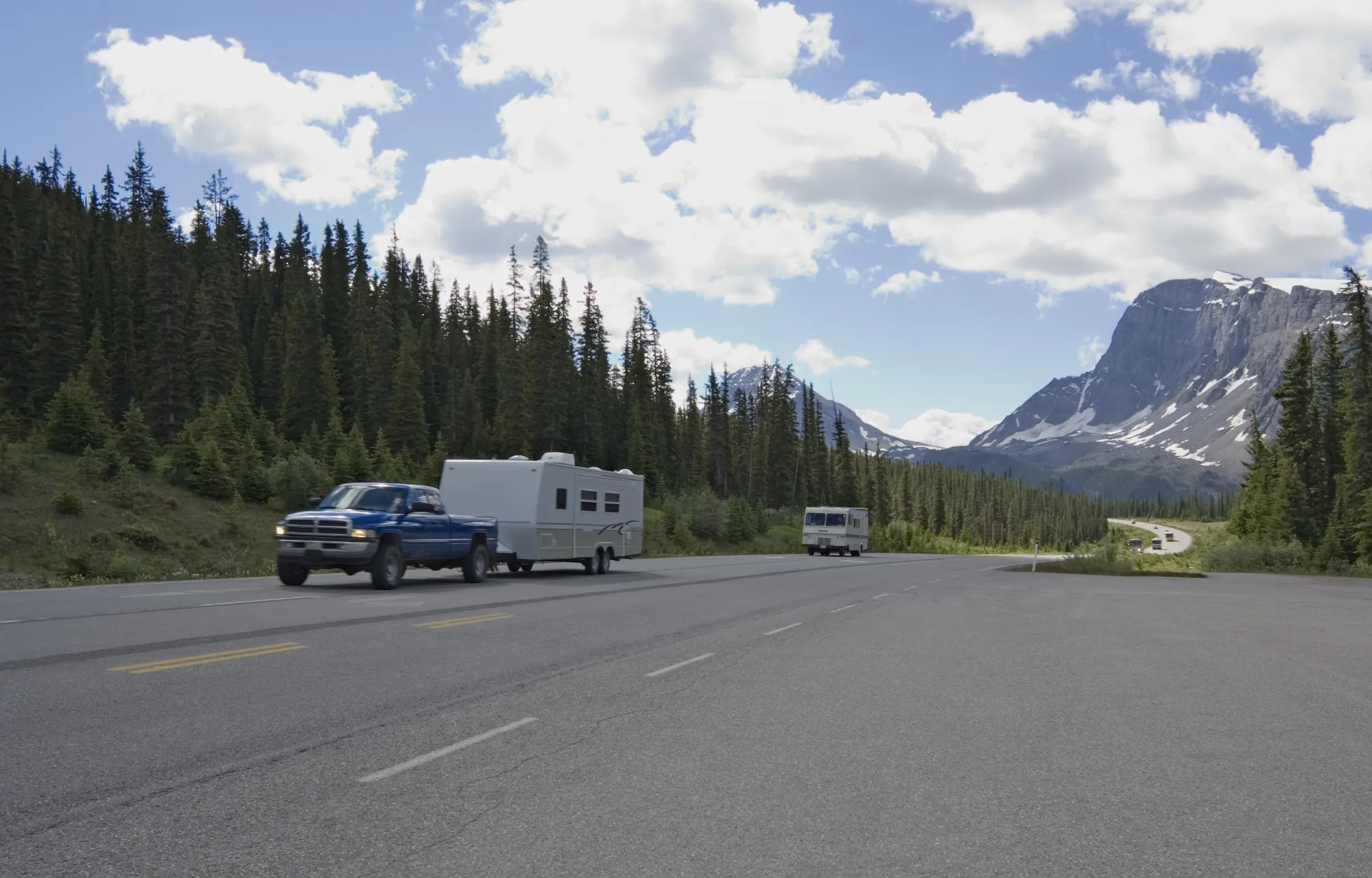A blue pickup truck towing a white travel trailer on a mountain highway lined with evergreen trees, with a motorhome ahead and snow-capped peaks in the distance.