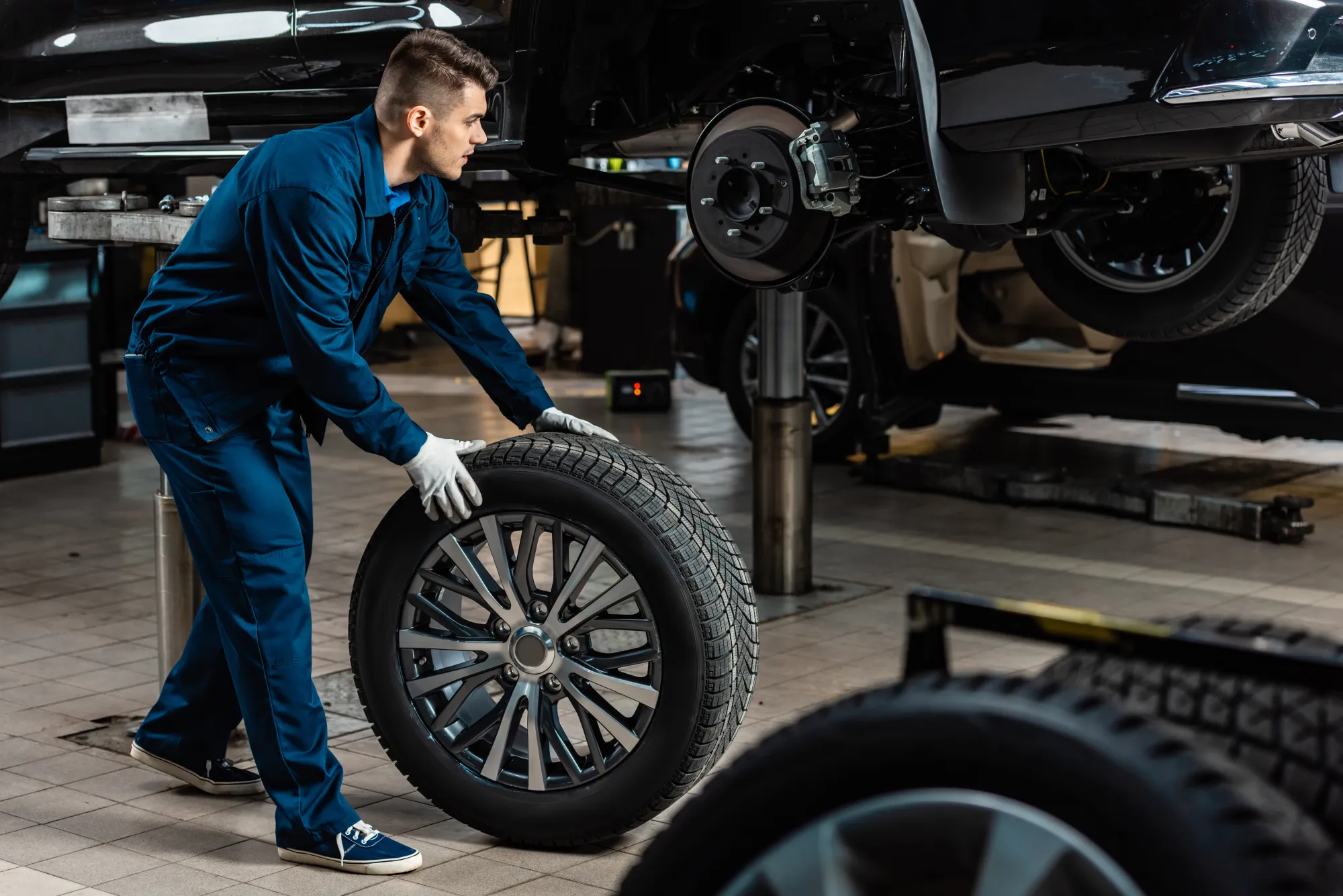 A mechanic in blue coveralls rolls a tire on an alloy wheel toward a vehicle raised on a shop lift with one wheel removed.