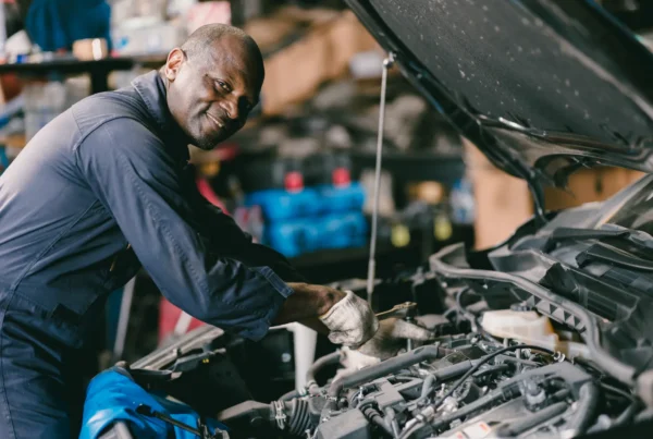 A smiling mechanic in navy coveralls works on a car engine under the open hood inside a repair shop.