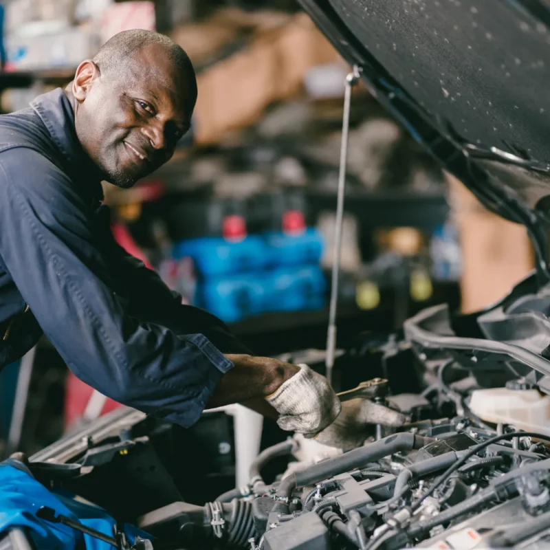 A smiling mechanic in navy coveralls works on a car engine under the open hood inside a repair shop.